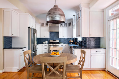 Transitional u-shaped medium tone wood floor and brown floor eat-in kitchen photo in Richmond with a farmhouse sink, recessed-panel cabinets, white cabinets, gray backsplash, stainless steel appliances and black countertops