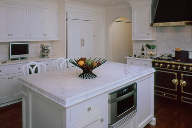 Enclosed kitchen - mid-sized traditional l-shaped dark wood floor enclosed kitchen idea in New York with a farmhouse sink, recessed-panel cabinets, white cabinets, marble countertops, white backsplash, stone slab backsplash, black appliances and an island