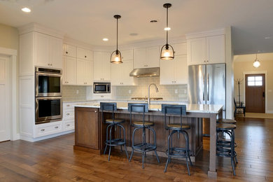 Open concept kitchen - mid-sized craftsman l-shaped medium tone wood floor and brown floor open concept kitchen idea in Other with an undermount sink, shaker cabinets, white cabinets, quartz countertops, beige backsplash, glass tile backsplash, stainless steel appliances and an island