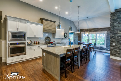 Example of a large arts and crafts single-wall medium tone wood floor and brown floor open concept kitchen design in Other with a farmhouse sink, shaker cabinets, white cabinets, granite countertops, white backsplash, stone tile backsplash, stainless steel appliances, an island and multicolored countertops