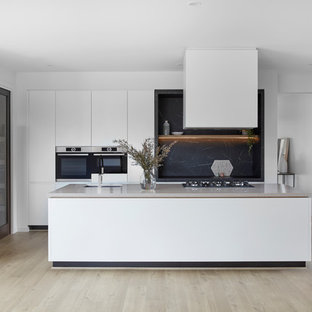 This is an example of a contemporary kitchen in Melbourne with an undermount sink, flat-panel cabinets, white cabinets, black splashback, stone slab splashback, stainless steel appliances, light hardwood floors, with island, beige floor and black benchtop.