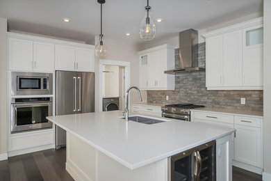 Example of a mid-sized transitional l-shaped dark wood floor and brown floor enclosed kitchen design in Calgary with a double-bowl sink, shaker cabinets, white cabinets, solid surface countertops, brown backsplash, stone tile backsplash, stainless steel appliances, an island and white countertops