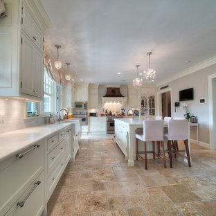 Photo of a large shabby-chic style l-shaped eat-in kitchen in Boston with a farmhouse sink, recessed-panel cabinets, beige cabinets, marble benchtops, white splashback, marble splashback, panelled appliances, travertine floors, with island, beige floor and white benchtop.
