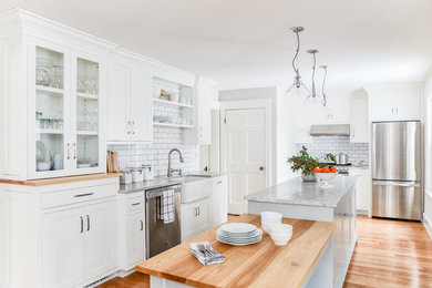 Example of a transitional l-shaped medium tone wood floor and brown floor kitchen design in Portland Maine with a farmhouse sink, shaker cabinets, white cabinets, white backsplash, subway tile backsplash, stainless steel appliances, an island and gray countertops