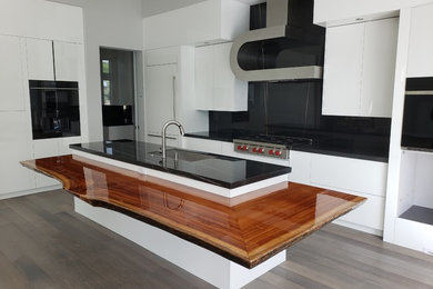 Example of a mid-sized minimalist l-shaped medium tone wood floor and gray floor open concept kitchen design in Tampa with flat-panel cabinets, white cabinets, soapstone countertops, black backsplash, stone slab backsplash, an island, black countertops, an undermount sink and paneled appliances