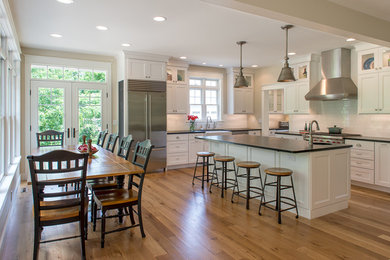 Example of a large transitional l-shaped light wood floor eat-in kitchen design in Boston with a farmhouse sink, recessed-panel cabinets, white cabinets, soapstone countertops, white backsplash, ceramic backsplash, stainless steel appliances and an island