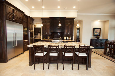 Large elegant l-shaped limestone floor open concept kitchen photo in Other with an undermount sink, raised-panel cabinets, dark wood cabinets, granite countertops, black backsplash, glass tile backsplash, stainless steel appliances and an island