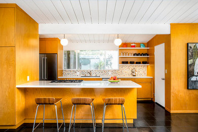 Example of a mid-sized 1950s galley black floor and slate floor open concept kitchen design in Los Angeles with an undermount sink, flat-panel cabinets, multicolored backsplash, stainless steel appliances, a peninsula, white countertops, medium tone wood cabinets, mosaic tile backsplash and quartz countertops