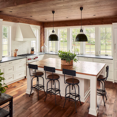 Example of a cottage l-shaped dark wood floor and brown floor kitchen design in Milwaukee with a farmhouse sink, shaker cabinets, white cabinets, paneled appliances, an island and black countertops