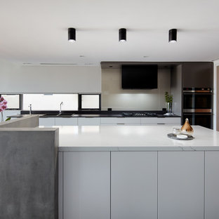 Photo of a contemporary galley eat-in kitchen in Sydney with a drop-in sink, flat-panel cabinets, white cabinets, grey splashback, glass sheet splashback, black appliances, with island, grey floor and white benchtop.
