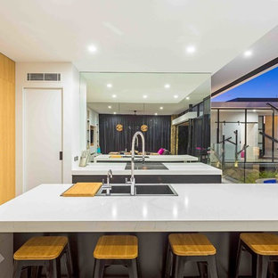Inspiration for a contemporary galley open plan kitchen in Gold Coast - Tweed with a drop-in sink, flat-panel cabinets, metallic splashback, mirror splashback, black appliances, concrete floors, with island, grey floor, light wood cabinets, granite benchtops and white benchtop.