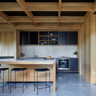 Photo of a midcentury kitchen in Melbourne with black cabinets, stainless steel benchtops, white splashback, ceramic splashback, black appliances, concrete floors, with island, grey floor, an integrated sink and flat-panel cabinets.