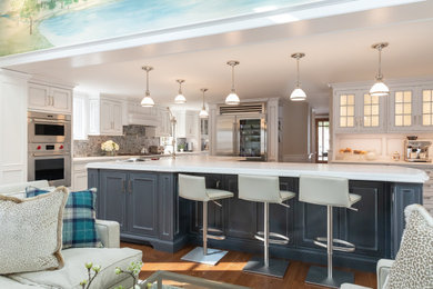 Example of a transitional medium tone wood floor and brown floor open concept kitchen design in New York with a farmhouse sink, beaded inset cabinets, blue cabinets, quartz countertops, gray backsplash, glass tile backsplash, stainless steel appliances, an island and white countertops