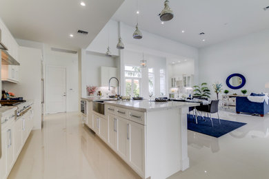 Large minimalist galley porcelain tile and beige floor open concept kitchen photo in Miami with a farmhouse sink, shaker cabinets, white cabinets, quartz countertops, white backsplash, stainless steel appliances, an island and stone slab backsplash