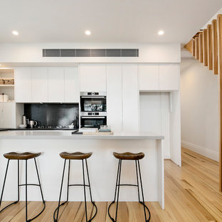 Photo of a contemporary l-shaped open plan kitchen in Sydney with black splashback, medium hardwood floors, with island, a drop-in sink, flat-panel cabinets, white cabinets, mosaic tile splashback and stainless steel appliances.