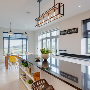 Photo of a large contemporary u-shaped eat-in kitchen in Devon with a farmhouse sink, flat-panel cabinets, white cabinets, granite benchtops, black splashback, stone slab splashback, black appliances, limestone floors, with island and beige floor.