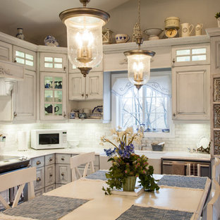 Ejemplo de cocina comedor en L, romántica, de tamaño medio, con fregadero sobremueble, armarios con paneles con relieve, puertas de armario con efecto envejecido, salpicadero blanco, salpicadero de azulejos tipo metro, electrodomésticos de acero inoxidable, suelo de madera clara, una isla y suelo marrón