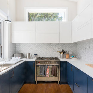 Design ideas for a transitional u-shaped kitchen in Sydney with a double-bowl sink, shaker cabinets, blue cabinets, white splashback, mosaic tile splashback, stainless steel appliances, medium hardwood floors, no island, brown floor and white benchtop.