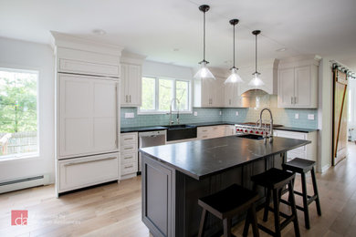 Open concept kitchen - large transitional l-shaped light wood floor and brown floor open concept kitchen idea in New York with a farmhouse sink, shaker cabinets, white cabinets, quartz countertops, green backsplash, subway tile backsplash, paneled appliances, an island and black countertops