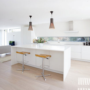 This is an example of a modern galley kitchen in Surrey with flat-panel cabinets, white cabinets, black appliances, light hardwood flooring, an island, beige floors and white worktops.