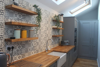 Design ideas for a medium sized contemporary galley kitchen/diner in Devon with a belfast sink, shaker cabinets, grey cabinets, wood worktops, grey splashback, ceramic splashback, light hardwood flooring and an island.