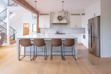 Example of a mid-sized transitional l-shaped light wood floor eat-in kitchen design in Calgary with a farmhouse sink, shaker cabinets, white cabinets, quartz countertops, white backsplash, ceramic backsplash, stainless steel appliances, an island and white countertops