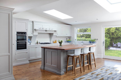 This is an example of a large classic l-shaped open plan kitchen in Dublin with a belfast sink, beaded cabinets, grey cabinets, quartz worktops, grey splashback, stone slab splashback, stainless steel appliances, medium hardwood flooring, an island and brown floors.