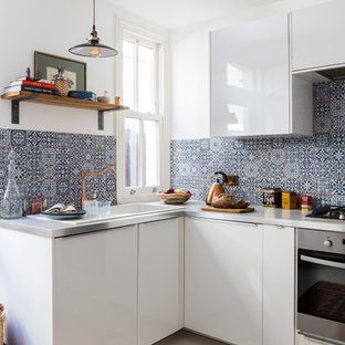 Small eclectic u-shaped separate kitchen in London with a drop-in sink, flat-panel cabinets, white cabinets, black splashback, ceramic splashback, stainless steel appliances, ceramic floors, no island and stainless steel benchtops.
