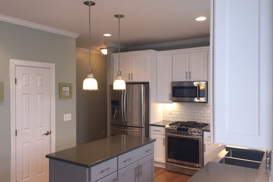 Example of a mid-sized transitional l-shaped light wood floor eat-in kitchen design in Raleigh with a farmhouse sink, recessed-panel cabinets, white cabinets, quartz countertops, white backsplash, subway tile backsplash, stainless steel appliances and an island