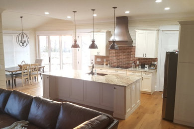 Example of a mid-sized arts and crafts l-shaped light wood floor and brown floor open concept kitchen design with recessed-panel cabinets, red backsplash, brick backsplash, an island, white cabinets, quartz countertops, stainless steel appliances and a farmhouse sink