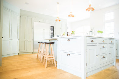 Inspiration for a large traditional u-shaped open plan kitchen in London with a submerged sink, shaker cabinets, green cabinets, composite countertops, white splashback, glass sheet splashback, stainless steel appliances, light hardwood flooring, an island and white worktops.