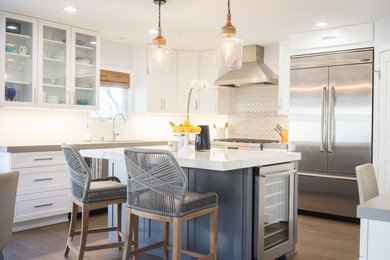 Photo of a classic l-shaped kitchen in Los Angeles with shaker cabinets, white cabinets, white splashback, stainless steel appliances, dark hardwood flooring, an island and brown floors.