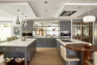 This is an example of an expansive industrial l-shaped open plan kitchen in Sussex with a submerged sink, shaker cabinets, grey cabinets, quartz worktops, glass sheet splashback, stainless steel appliances, light hardwood flooring, multiple islands, white worktops and a coffered ceiling.