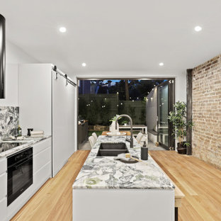 Photo of a contemporary galley kitchen in Sydney with an undermount sink, flat-panel cabinets, white cabinets, multi-coloured splashback, stone slab splashback, black appliances, light hardwood floors, with island, beige floor and multi-coloured benchtop.