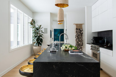 Example of a mid-sized trendy galley light wood floor and beige floor eat-in kitchen design in Chicago with flat-panel cabinets, white cabinets, marble countertops, black backsplash, ceramic backsplash, stainless steel appliances, an island and black countertops