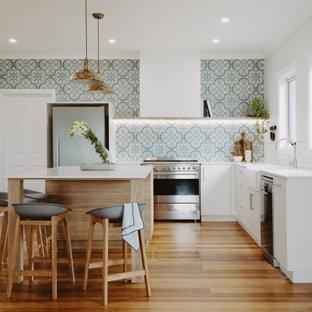 This is an example of a large contemporary l-shaped eat-in kitchen in Hobart with a farmhouse sink, white cabinets, granite benchtops, cement tile splashback, stainless steel appliances, light hardwood floors, with island, white benchtop, flat-panel cabinets, green splashback and brown floor.