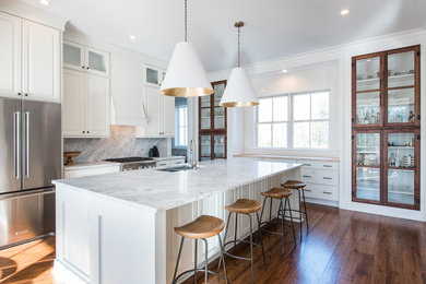 Inspiration for a large transitional l-shaped medium tone wood floor and brown floor open concept kitchen remodel in Charleston with an undermount sink, raised-panel cabinets, white cabinets, marble countertops, white backsplash, stone slab backsplash, stainless steel appliances and an island