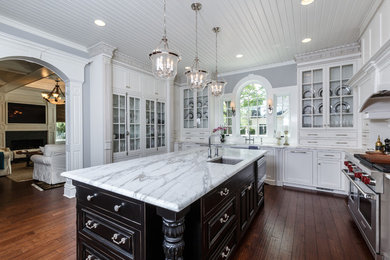 Example of a large ornate u-shaped medium tone wood floor enclosed kitchen design in Cincinnati with glass-front cabinets, white cabinets, marble countertops, an island, an undermount sink, white backsplash, subway tile backsplash and stainless steel appliances