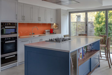 Traditional galley kitchen/diner in London with shaker cabinets, grey cabinets, orange splashback, black appliances, an island and grey floors.