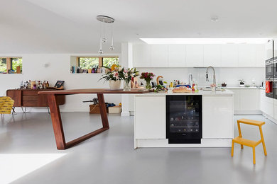 Design ideas for a contemporary galley open plan kitchen in London with a built-in sink, flat-panel cabinets, white cabinets, composite countertops, white splashback, ceramic splashback, black appliances, an island, grey floors and white worktops.