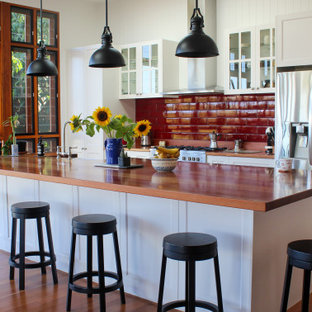 This is an example of a country galley kitchen in Brisbane with a farmhouse sink, shaker cabinets, white cabinets, wood benchtops, red splashback, subway tile splashback, stainless steel appliances, medium hardwood floors, with island, brown floor and brown benchtop.