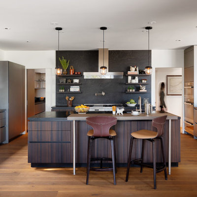Example of a large trendy u-shaped medium tone wood floor and brown floor kitchen design in Los Angeles with an island, an undermount sink, flat-panel cabinets, dark wood cabinets, black backsplash, stainless steel appliances and black countertops