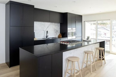 Example of a mid-sized minimalist single-wall light wood floor and beige floor eat-in kitchen design in Vancouver with an undermount sink, flat-panel cabinets, black cabinets, solid surface countertops, white backsplash, marble backsplash, paneled appliances, an island and black countertops