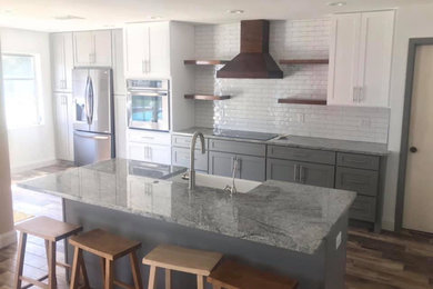 Modelo de cocina lineal de estilo de casa de campo de tamaño medio con fregadero sobremueble, armarios estilo shaker, puertas de armario grises, encimera de granito, salpicadero blanco, salpicadero de azulejos tipo metro, electrodomésticos de acero inoxidable, suelo de madera en tonos medios, una isla, suelo marrón y encimeras grises