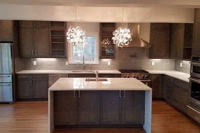 Large transitional l-shaped medium tone wood floor and brown floor kitchen photo in New York with an undermount sink, shaker cabinets, dark wood cabinets, gray backsplash, stainless steel appliances and an island