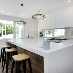 Beach style l-shaped eat-in kitchen in Sunshine Coast with an undermount sink, flat-panel cabinets, white cabinets, mirror splashback, stainless steel appliances, medium hardwood floors, with island, brown floor and white benchtop.