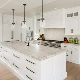 Expansive country single-wall open plan kitchen in Salt Lake City with a farmhouse sink, beaded inset cabinets, white cabinets, quartzite benchtops, beige splashback, limestone splashback, white appliances, light hardwood floors, multiple islands, beige floor and beige benchtop.