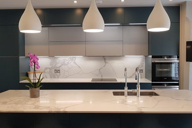 This is an example of a large contemporary open plan kitchen in London with a single-bowl sink, flat-panel cabinets, green cabinets, quartz worktops, white splashback, stone slab splashback, stainless steel appliances, light hardwood flooring, an island, grey floors and white worktops.