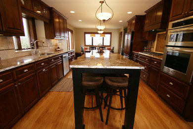 This is an example of a large classic galley kitchen in Minneapolis with a submerged sink, raised-panel cabinets, dark wood cabinets, granite worktops, beige splashback, ceramic splashback, stainless steel appliances, medium hardwood flooring and an island.