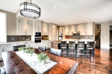 Large transitional l-shaped medium tone wood floor and brown floor eat-in kitchen photo in Calgary with a farmhouse sink, recessed-panel cabinets, white cabinets, quartzite countertops, gray backsplash, glass tile backsplash, paneled appliances and an island